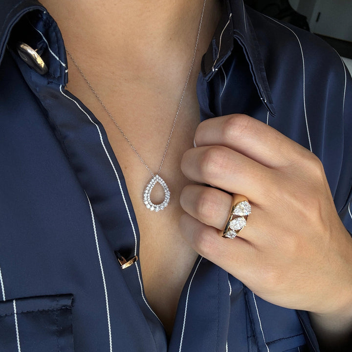 a white gold teardrop-shaped pendant necklace with a halo setting, paired with a gold three stone diamond ring on their finger