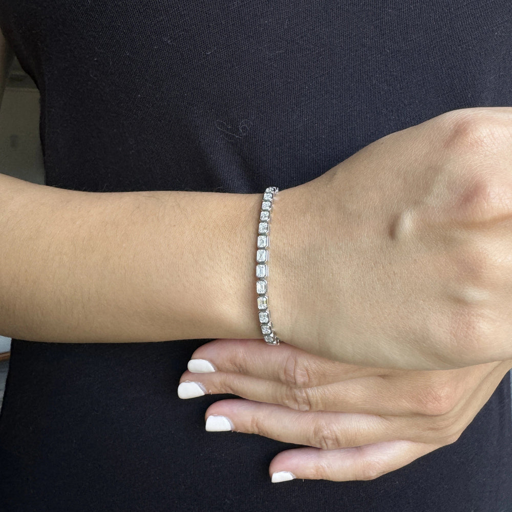 A close-up of a person's wrist wearing a white gold tennis bracelet with emerald cut diamonds, set in an east west setting