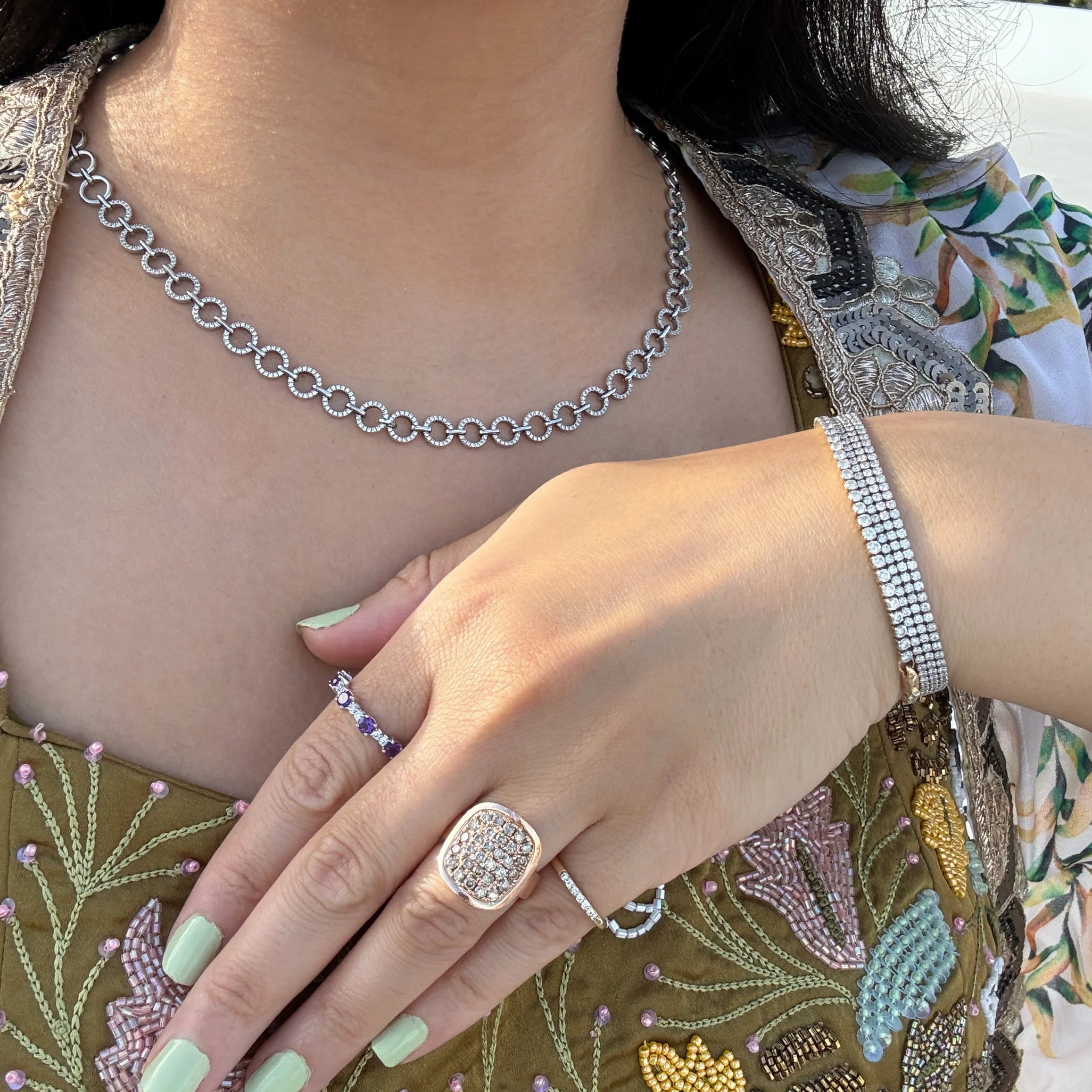 A close-up lifestyle shot of a person wearing a white gold diamond necklace, stacked diamond tennis bracelets, and gold rings, including a prominent square-cut ring with a pavé diamond setting, an amethyst and diamond band ring, and a pave diamond pinky ring.