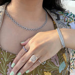 A close-up lifestyle shot of a person wearing a white gold diamond necklace, stacked diamond tennis bracelets, and gold rings, including a prominent square-cut ring with a pavé diamond setting, an amethyst and diamond band ring, and a pave diamond pinky ring.