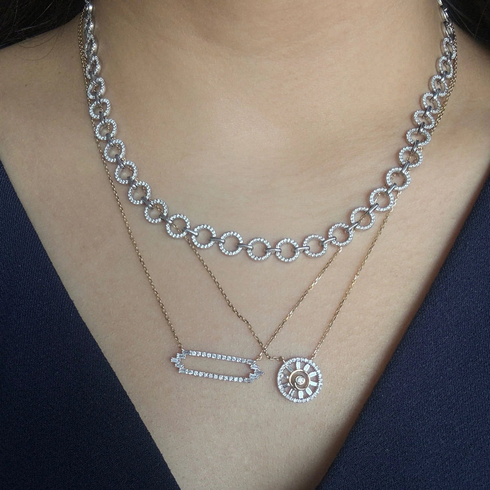 A close-up of a model wearing layering necklaces. The diamond necklaces include a white gold collar necklace with a repeating circular link pattern, a yellow gold diamond necklace with pave diamonds and baguette diamonds, and a matching yellow gold chain with a circular sunburst design also made of pave diamond and baguette diamonds.