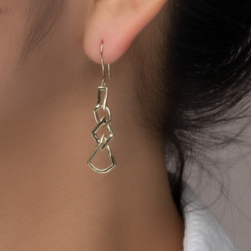 unique jewelry, unique earrings, fashion jewelry, fashion earrings, drop earrings in yellow gold, dangling earrings, dangling drop earrings with a design of geometric shapes intertwined with each other. Earrings have a fishhook back. Earrings on model.
